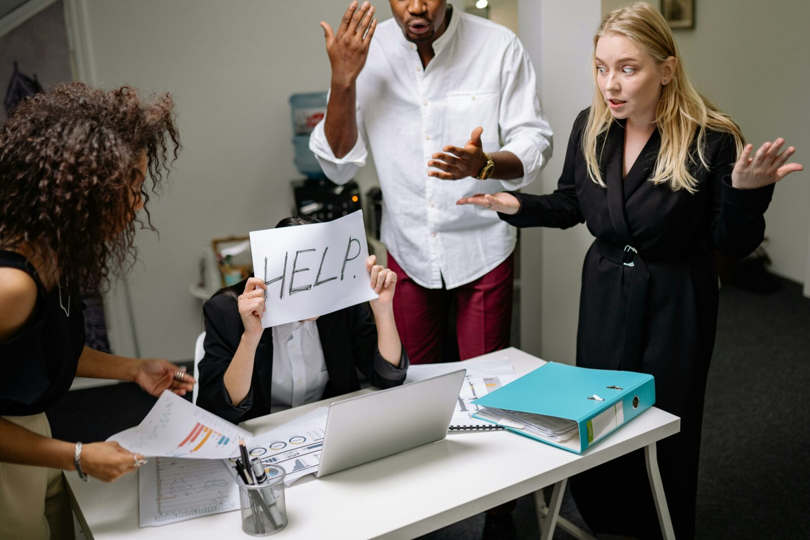 Une personne assise au bureau tenant une feuille avec l'inscription "HELP" entourée de trois autres personnes montrant des signes d'énervement.