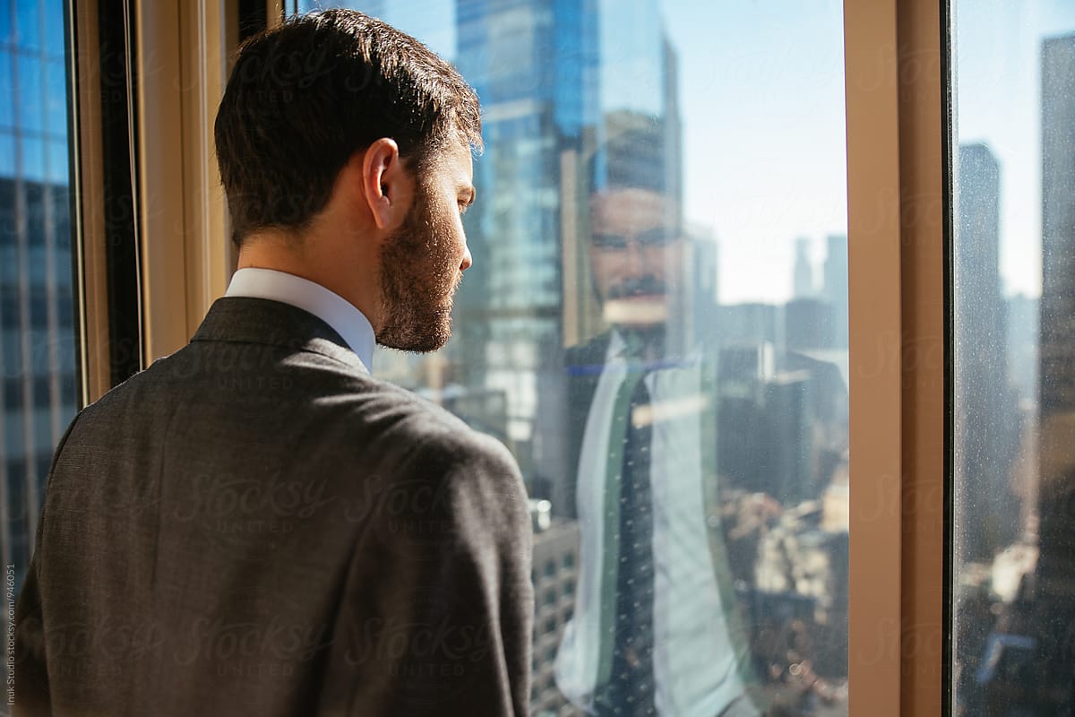 Homme en costume observant la ville depuis une fenêtre, illustrant la posture professionnelle et la prise de recul en milieu de travail.